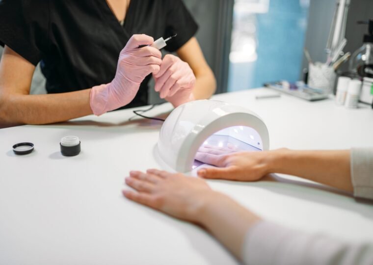 Client's hand under a white UV nail lamp while a pink-gloved technician holds a small brush at a white table.