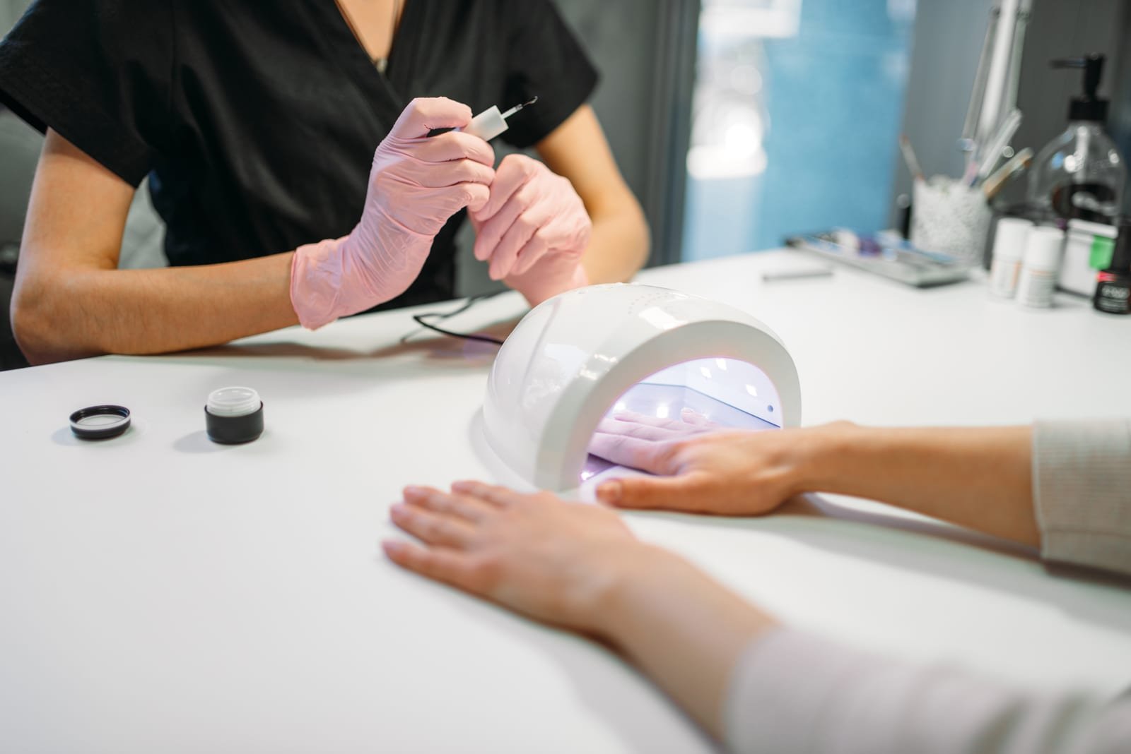 Client's hand under a white UV nail lamp while a pink-gloved technician holds a small brush at a white table.