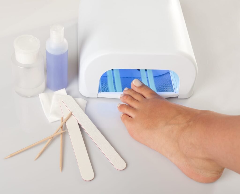 A foot rests with its toes under the blue light of a white UV nail lamp, with nail files, wooden cuticle sticks, cotton pads and two small bottles of liquid arranged on the white surface…