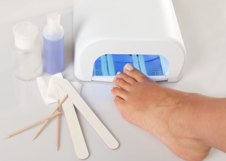 A foot rests with its toes under the blue light of a white UV nail lamp, with nail files, wooden cuticle sticks, cotton pads and two small bottles of liquid arranged on the white surface…