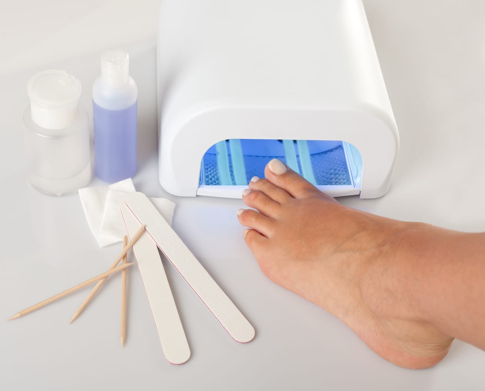 A foot rests with its toes under the blue light of a white UV nail lamp, with nail files, wooden cuticle sticks, cotton pads and two small bottles of liquid arranged on the white surface…